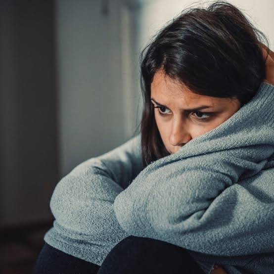 A woman sitting alone, arms around her knees, looking quietly heartbroken